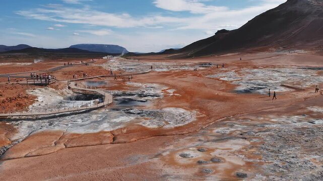 The Hverir geothermal area in Iceland features steaming fumaroles, bubbling mud pools, mineral streaked ground, and visitors on a boardwalk.