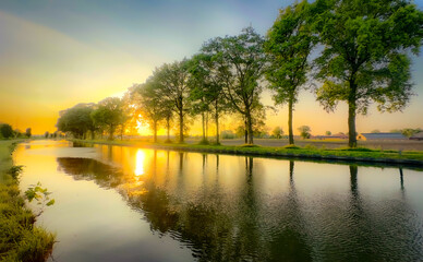 A serene sunset casts warm colors and reflections on a calm canal, embraced by lush trees and...
