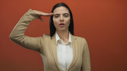 Hispanic woman in a beige suit performs a military salute against a bold red wall background,...