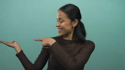 Obraz premium Smiling hispanic woman points to her palm against a green backdrop, exuding confidence and charm with a friendly gesture.