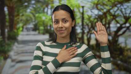 Hispanic woman making a pledge gesture in a green park with sunlight filtering through the trees,...