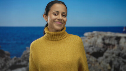 Young woman smiling joyfully in a mustard sweater by the serene seaside, capturing a carefree and...