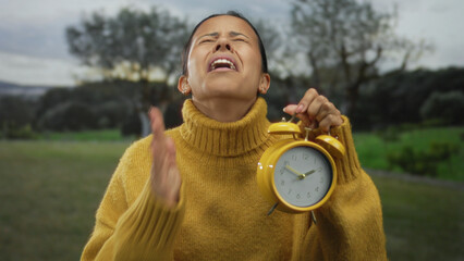 Woman outdoors holding yellow alarm clock in a green park expressing frustration with lush trees in...