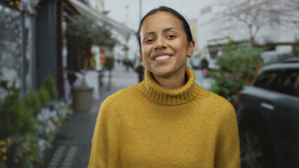 Woman smiling outdoors on a city street, wearing a yellow sweater and gesturing with hands,...