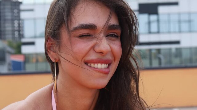 A young woman with long hair relaxes on a rooftop, enjoying the warm weather. She smiles as the wind gently blows her hair. The girl is winking seductively.