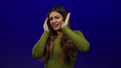 Hispanic woman with long hair covers her face with painted nails against a blue background,...