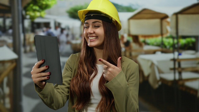 Woman smiling on a terrace during a video call, wearing a yellow hard hat and casual jacket, showcasing a blend of professional and relaxed outdoor ambiance.
