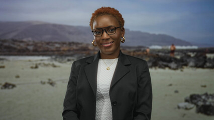 Woman wearing glasses and a necklace smiles with eyes closed on rocky beach shore under soft...