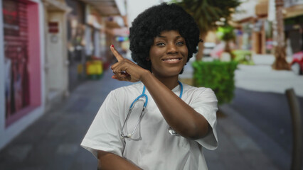 Young african american woman doctor with stethoscope points finger on city street lined with shops;...