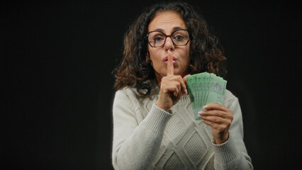 Middle-aged woman in glasses holding malaysian ringgit banknotes with a gesture for silence against...