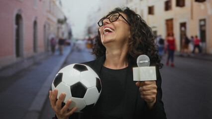 Woman reporter holding soccer ball and microphone on a lively street outdoors, capturing the...
