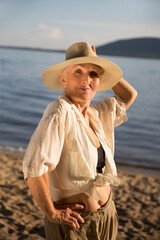 smiling elderly woman wearing a straw hat enjoys the sun on the seashore or river in the summer evening. nature walks.  spends time in nature in summer.  