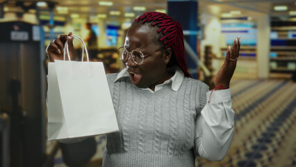 Woman wearing glasses and sweater excitedly holds shopping bag indoors with blurred background...