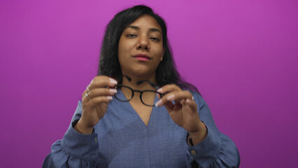 African american woman removes black framed glasses using both hands in well lit purple studio;...