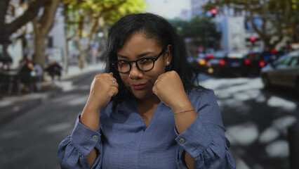 Woman wearing glasses and blue shirt with raised fists in boxing stance on city street crosswalk;...