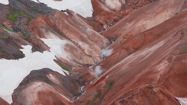 Aerial view of Kerlingarfjoll and Studlagil Canyon - vibrant red and orange hills with geothermal steam, patches of snow, and flowing streams in a rugged Icelandic terrain.