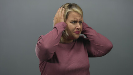 Young blonde woman pressing hands against ears and grimacing in a grey studio setting; discomfort.