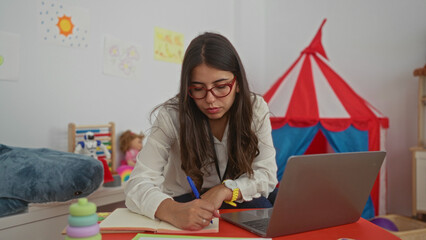 Young woman teacher in a colorful kindergarten taking notes with a laptop on a table surrounded by...