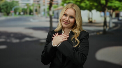 Woman expressing gratitude outdoors in city setting with trees and buildings, blonde hair, hands on...
