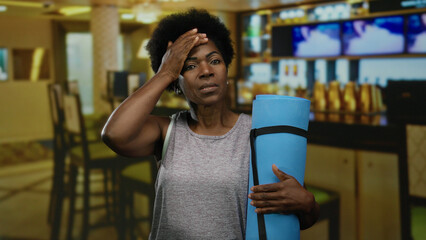 Woman in restaurant holds yoga mat, appearing stressed with hand on forehead, showcasing an african american in a coffee shop setting for a relatable lifestyle image.