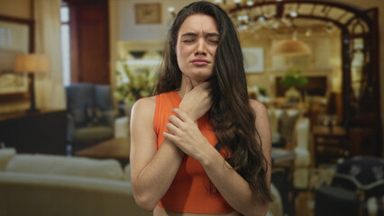 Young hispanic woman clutching her throat inside a large hotel building interior while wincing in...