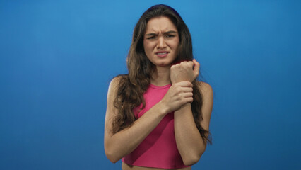 Teenage hispanic woman holds wrist with furrowed brow showing pain gesture in blue studio; distress.