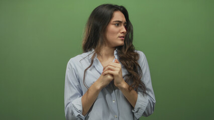 Young hispanic woman clasping hands and furrowing brows with tense posture in green studio;...