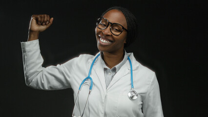 Woman in white coat with stethoscope raising fist confidently against dark background showing...