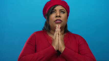 Woman praying wearing red beret and sweater, standing against a vibrant blue background, expressing...
