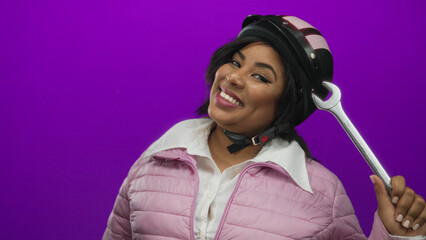 Woman wearing helmet holds wrench smiling against purple background, showcasing young confident latin plus size female in pink jacket.