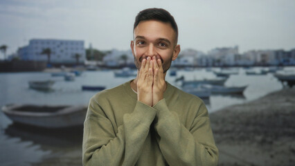 Young hispanic man covering mouth in surprise against seaside promenade with boats in the...