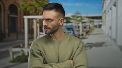 Obraz premium Young man with beard standing on urban street with thoughtful expression wearing casual sweater under blue sky in busy city background.