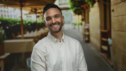 Obraz premium Young man smiling playfully outdoors in urban street setting with blurred background of tables and buildings, capturing joyful and lively city vibe during daytime.
