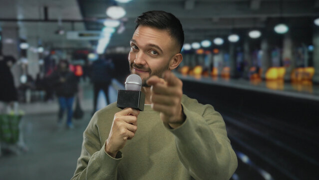 Young man holding microphone in underground station giving interview with blurred commuters walking in background, a hispanic adult in casual attire standing confidently.