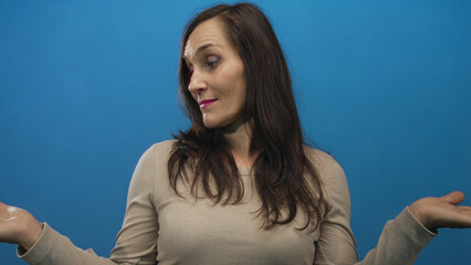 Woman shrugging with uncertainty against a vivid blue background wall showing a casual and confused...