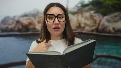 Woman reading book and adjusting glasses at rocky seaside beach with turquoise water behind her and...
