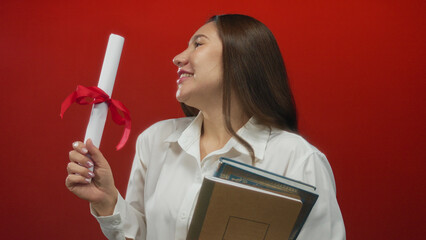 Young hispanic woman holds diploma with red ribbon and stack of books in studio; proud achievement.