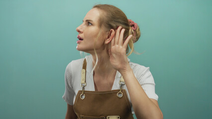 Young blonde woman in an apron gestures listening against an isolated green background, expressing...