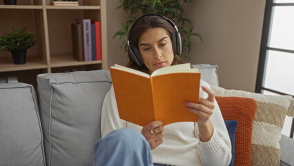 Woman reading book on couch wearing headphones in cozy living room with bookshelves and pillows,...
