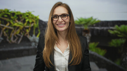 Young caucasian woman wearing glasses stands in a leafy park beside a stone building wall with...