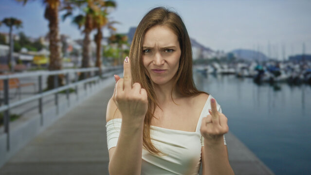 Woman holds up two middle fingers at a port dock under bright daylight, scowling intensely at the lens; defiance.