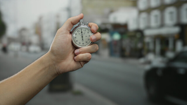 Hand holding a stopwatch in an urban street setting with blurred town buildings, symbolizing time management and precision by a man in an outdoor city environment.