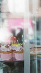Colorful desserts displayed in a glass case include cupcakes with pink frosting in a bustling cafeteria or bakery setting, offering a vibrant and enticing atmosphere for patrons.