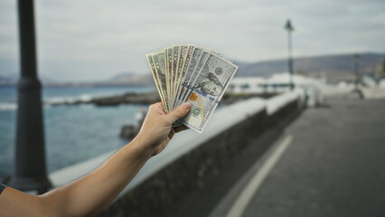 Man holding american dollars in hand by seaside, showcasing currencies with blurred ocean in...
