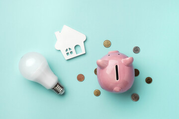 A pink piggy bank surrounded by coins with a light bulb and house model on light blue background...