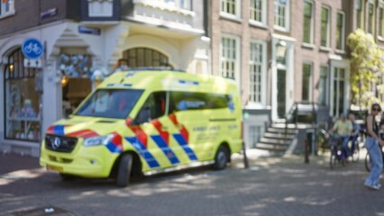 Bokeh image of a bright yellow ambulance parked on a cobblestone street, with defocused pedestrians...