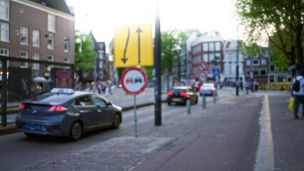 Blurred european city street scene featuring cars and people with bokeh effect in a defocused setting during daytime among urban buildings and traffic signs.