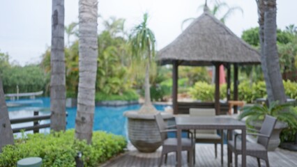 Blurred view of a luxury tropical resort featuring a bokeh scene with outdoor seating beside a sparkling pool surrounded by lush greenery and palm trees under sunny skies.