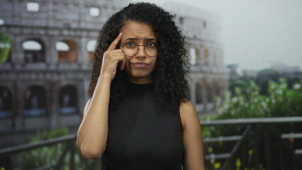 Fototapeta premium Young hispanic woman finger to head gesture by roman colosseum building outdoors in daylight on a street balcony; curiosity.