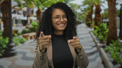 Young hispanic woman points finger to camera on a sunny park walkway lined with palm trees;...